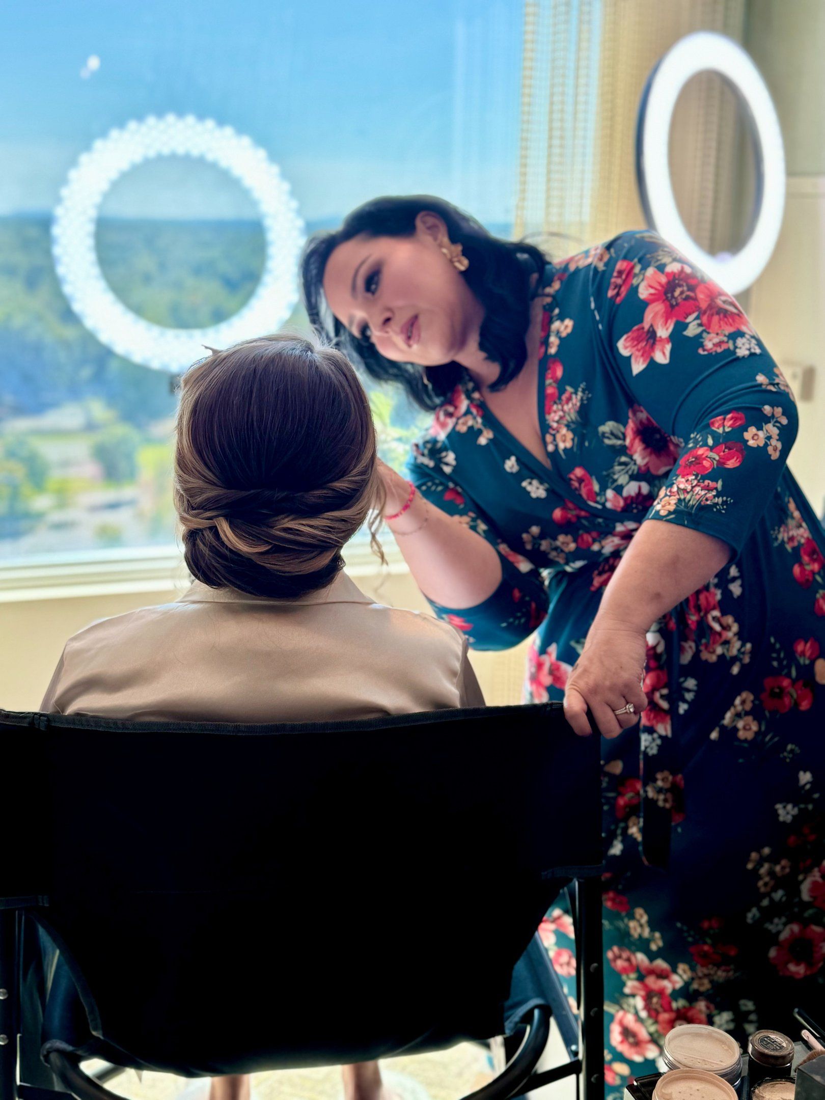 Makeup artist refines the bride’s makeup beside a window; twisted low updo and ring light showcase elegant wedding hair.