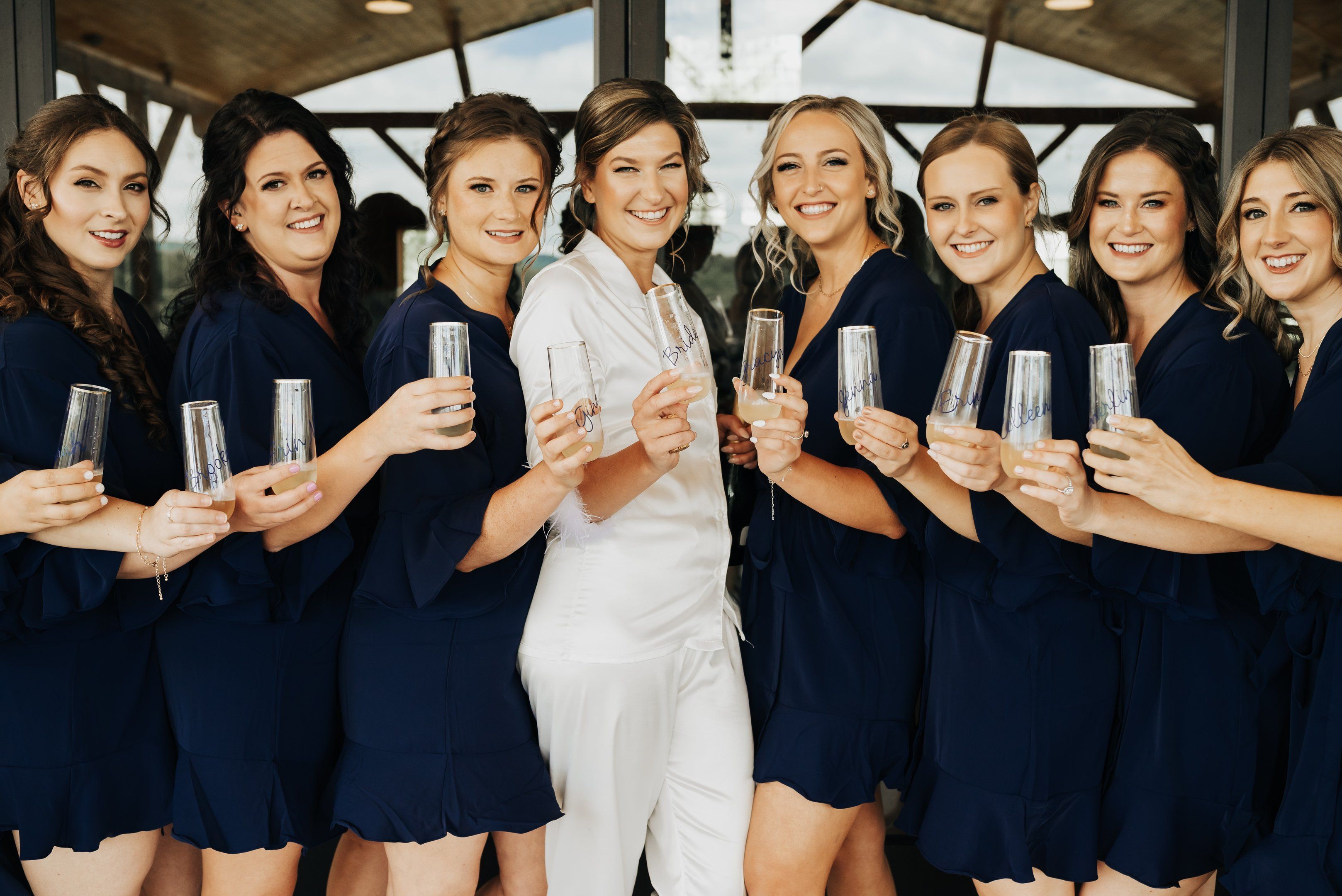 Bride in white pjs with bridesmaids in navy robes holding champagne glasses, showing elegant wedding hair and natural makeup.