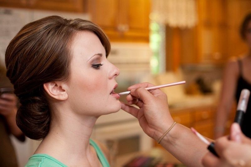 Bride with elegant updo receives wedding makeup touch as artist applies lipstick in a candid getting ready moment.