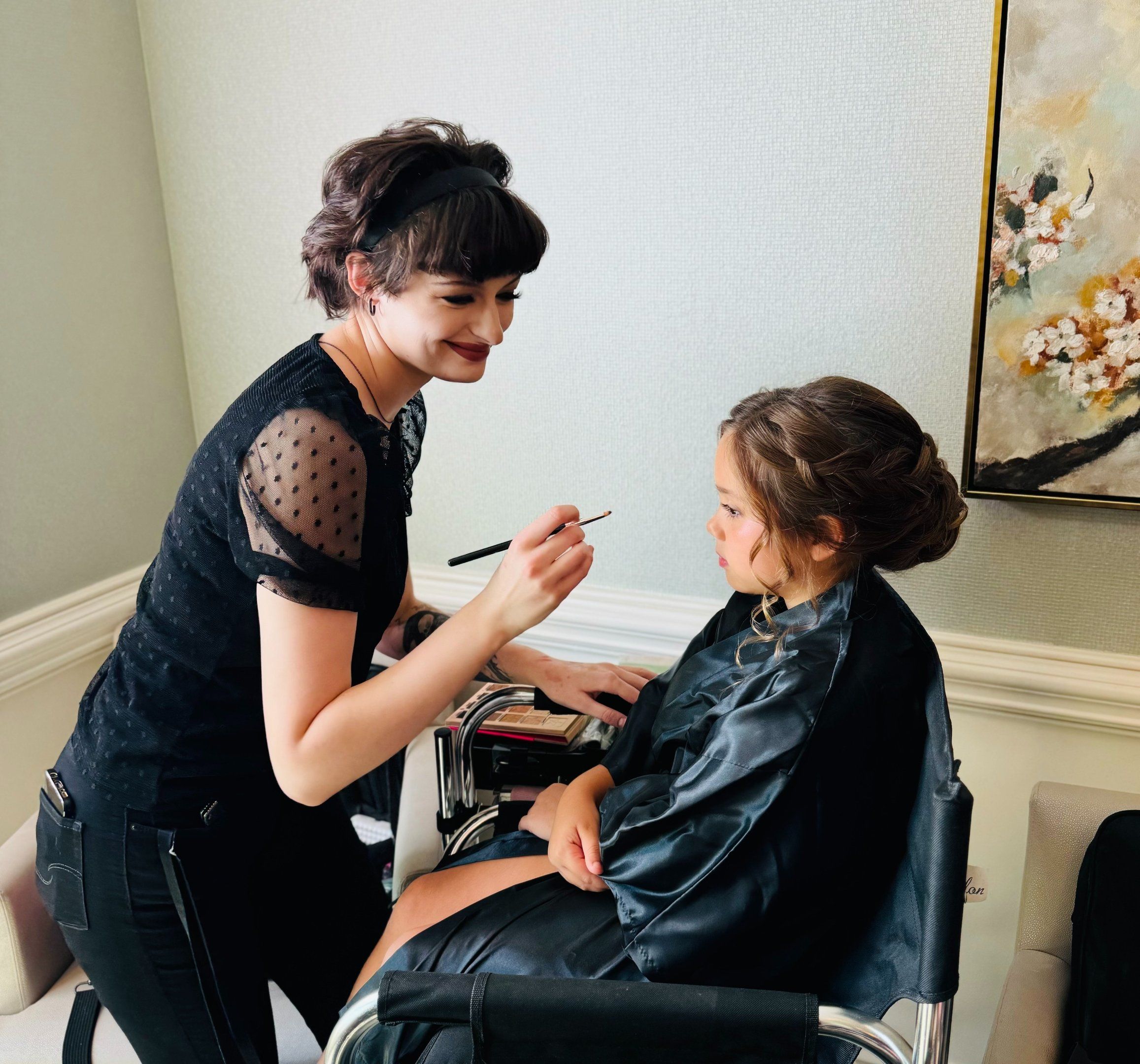 Makeup artist adds blush to a flower girl with a braided updo, capturing sweet wedding hair and makeup getting ready moment.