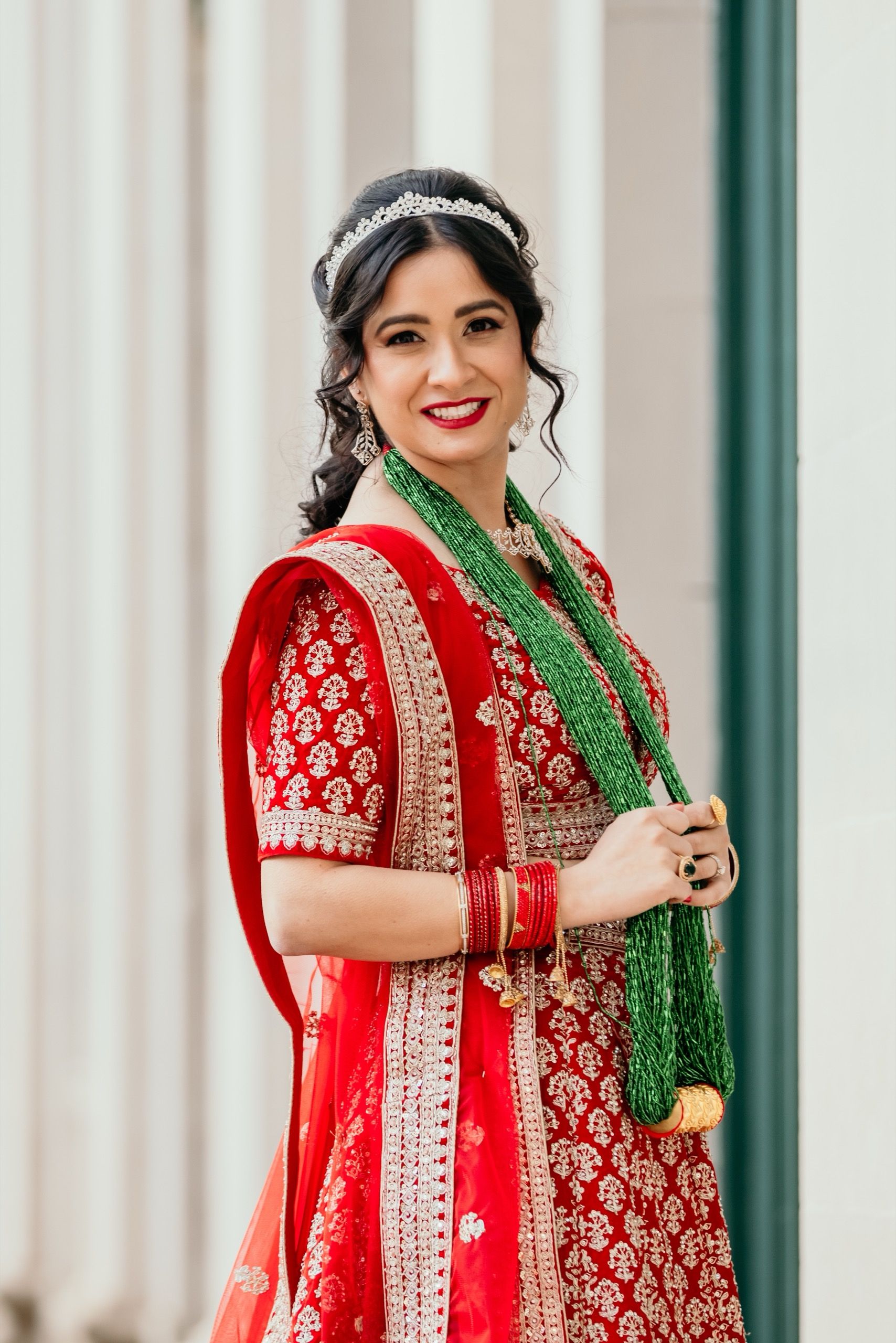 Indian bride with bridal makeup wearing red lehenga, green necklace, tiara, and bold red lipstick for traditional wedding.