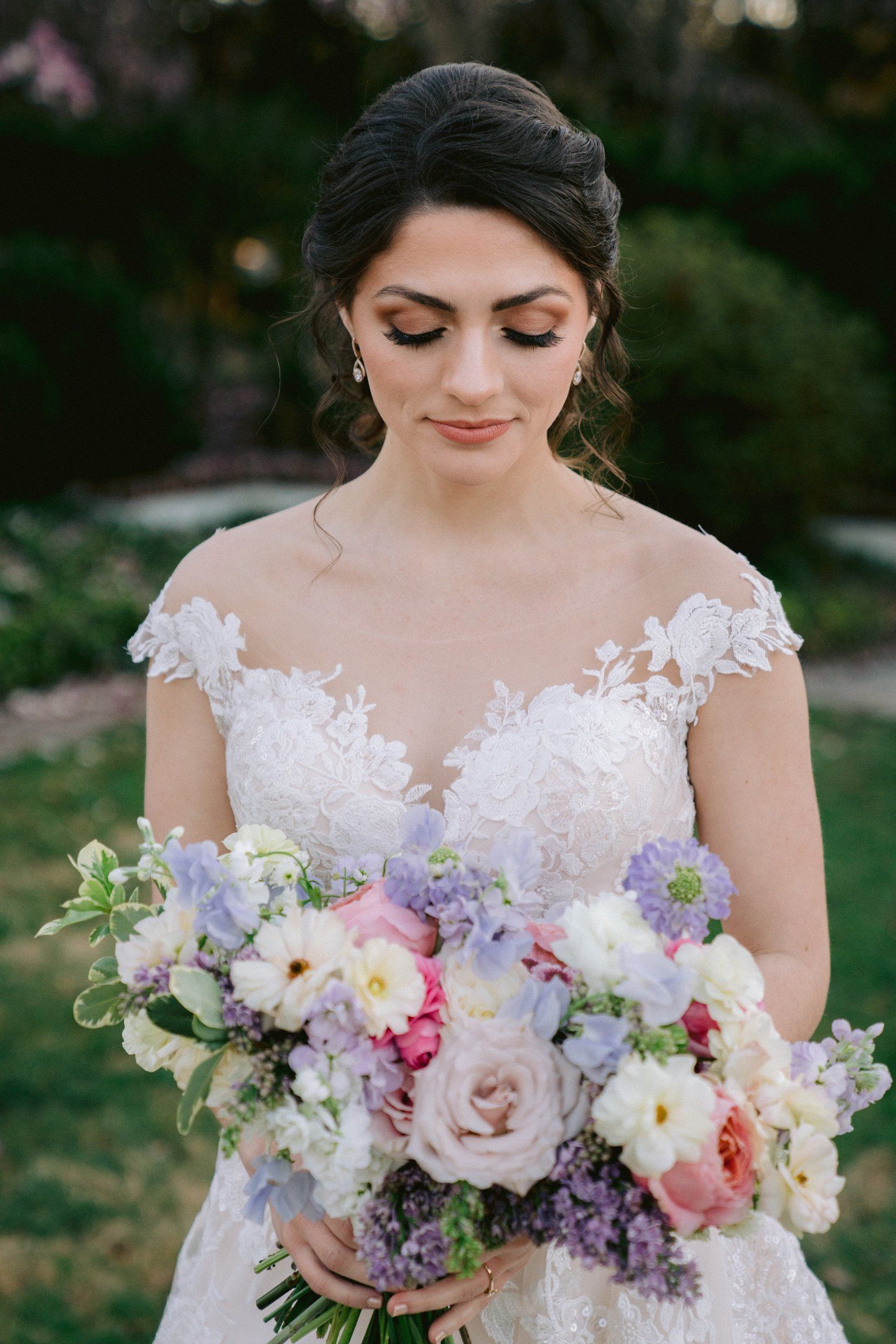 Bride with romantic bridal updo, soft wedding makeup, long lashes and neutral tones, holding pastel bouquet outdoors.